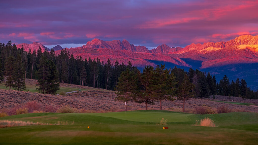 A vibrant sunset illuminates the rugged mountain peaks in the background with hues of purple and pink reflecting off scattered clouds. In the foreground, the golf course is in twilight shadow.