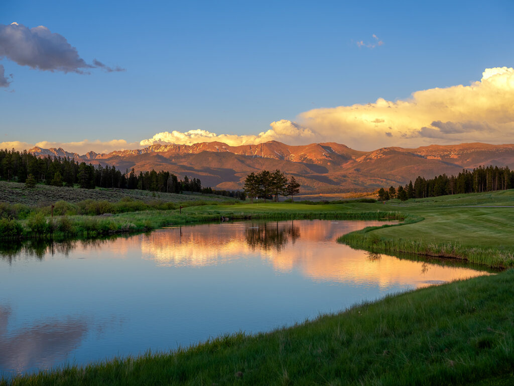 Alt text: "The golden hour sun casts a warm glow over a tranquil golf course with a reflective pond in the foreground.
