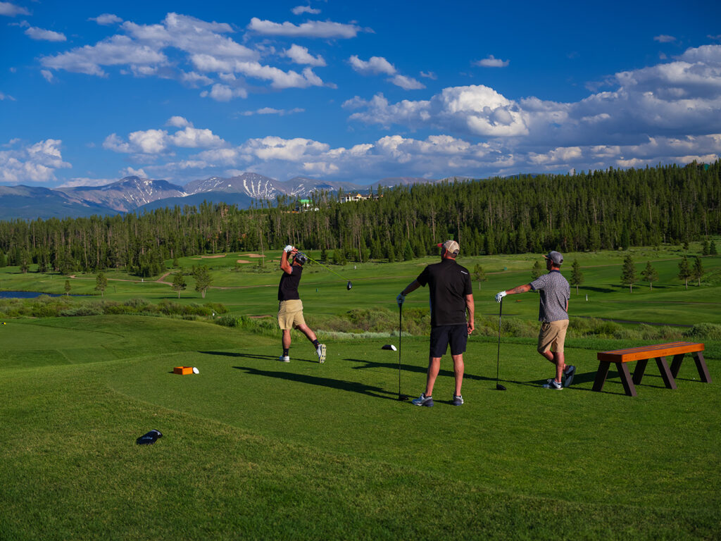 Three golfers on a tee box at Pole Creek Golf Club enjoying a sunny day on the course. One golfer is in mid-swing, while the other two watch the flight of the ball.