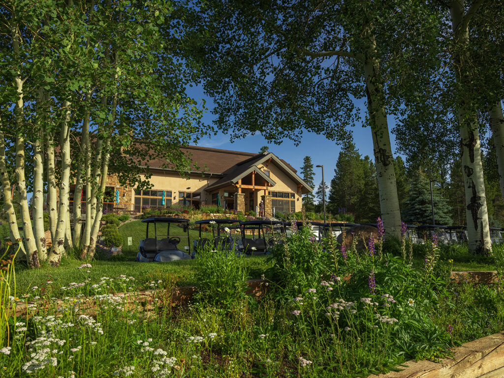 The clubhouse at Pole Creek Golf Club is framed by quaking aspen trees under a clear blue sky. A lush garden with wildflowers and well-tended landscaping leads to the rustic building adorned with a stone facade