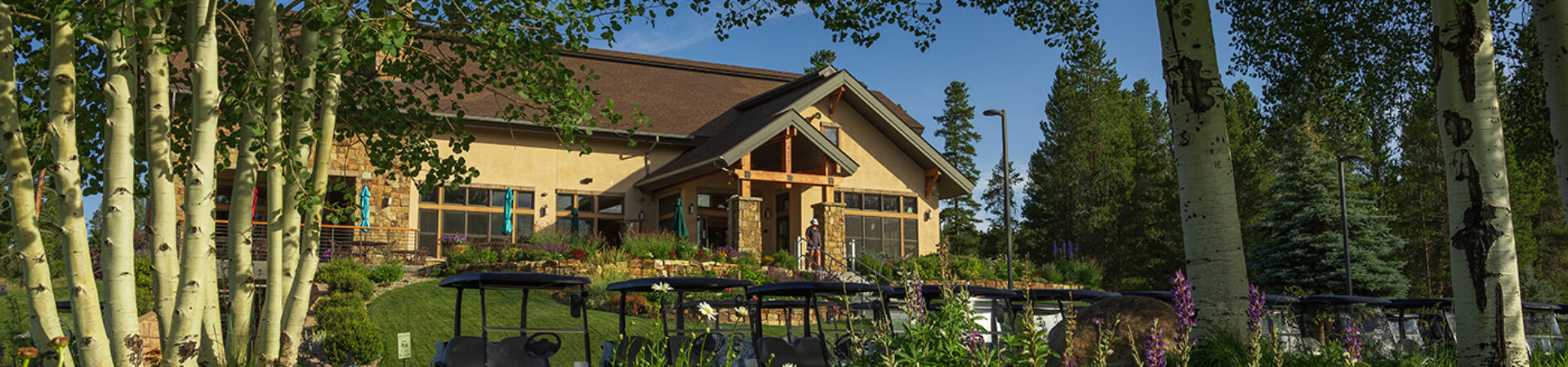 The clubhouse at Pole Creek Golf Club is framed by quaking aspen trees under a clear blue sky. A lush garden with wildflowers and well-tended landscaping leads to the rustic building adorned with a stone facade